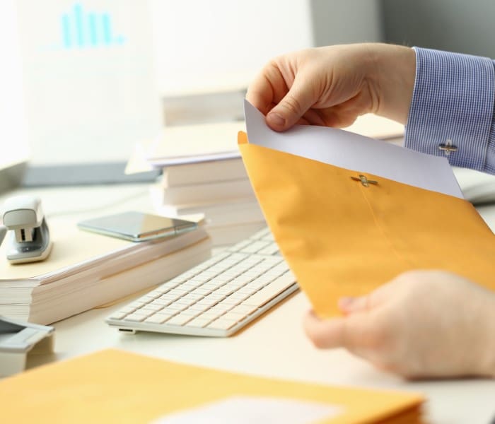 A person placing a document into a yellow envelope on a desk with a keyboard, phone, stapler, and stacked papers.