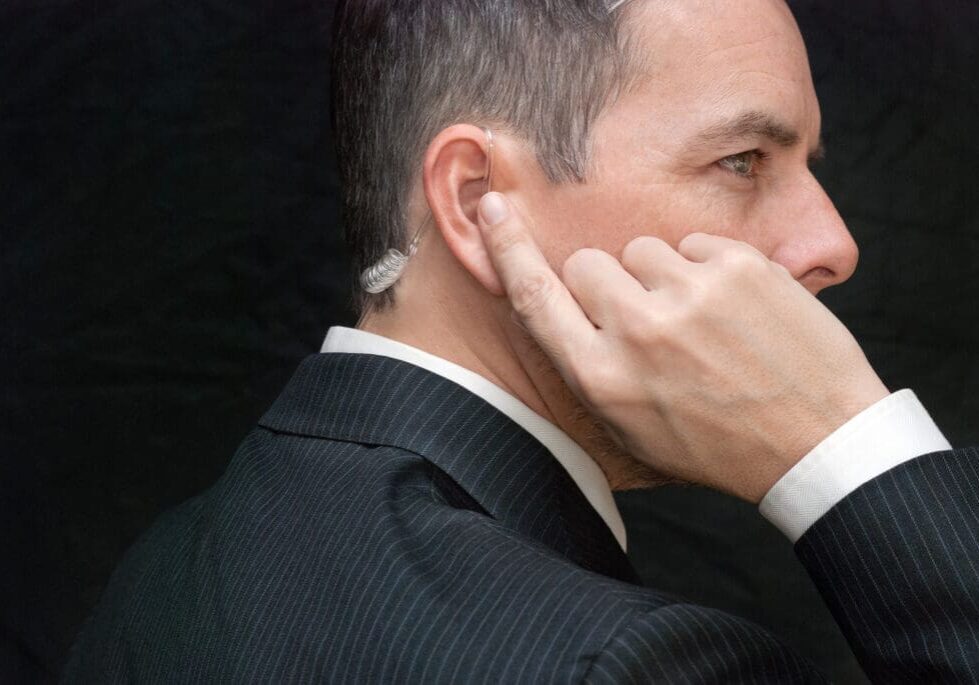 A man in a suit with an earpiece is holding his finger to his ear, standing against a dark background.