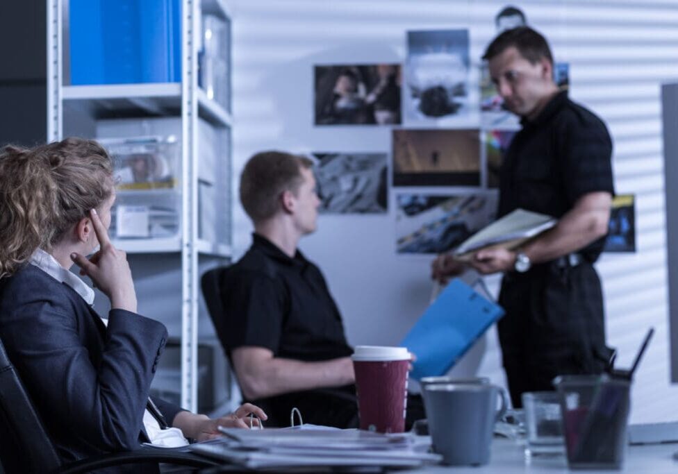 Three people in a dimly lit office, sitting or standing around a desk with documents, a laptop, and a coffee cup.