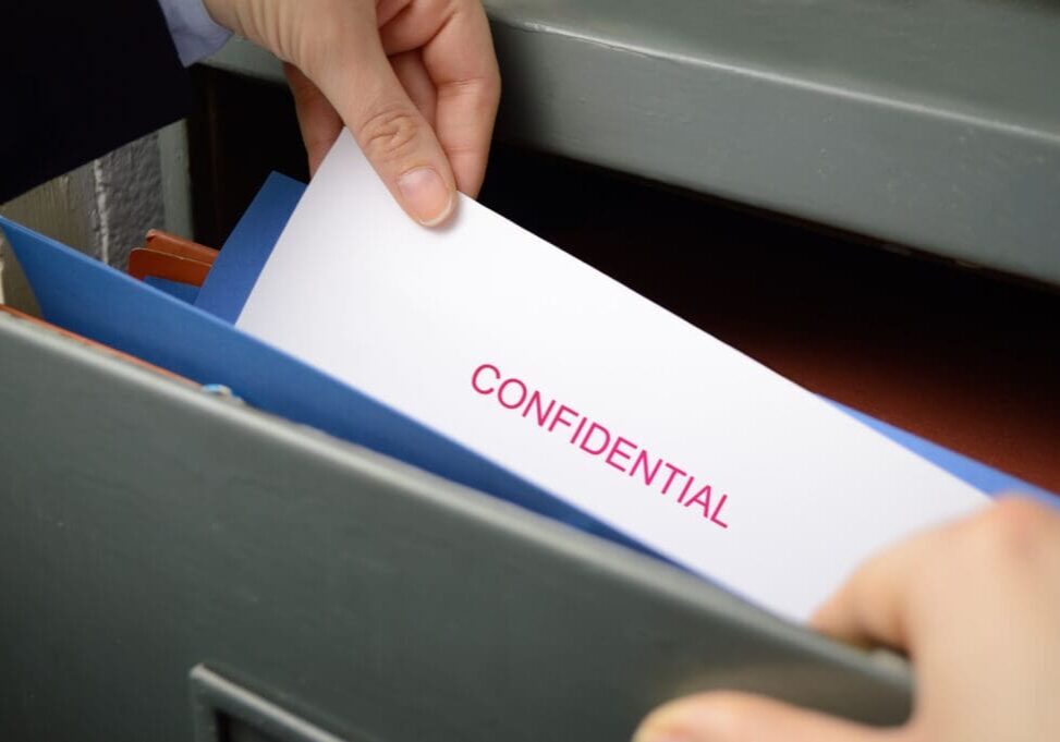 Hands placing a document labeled "Confidential" into a file drawer.